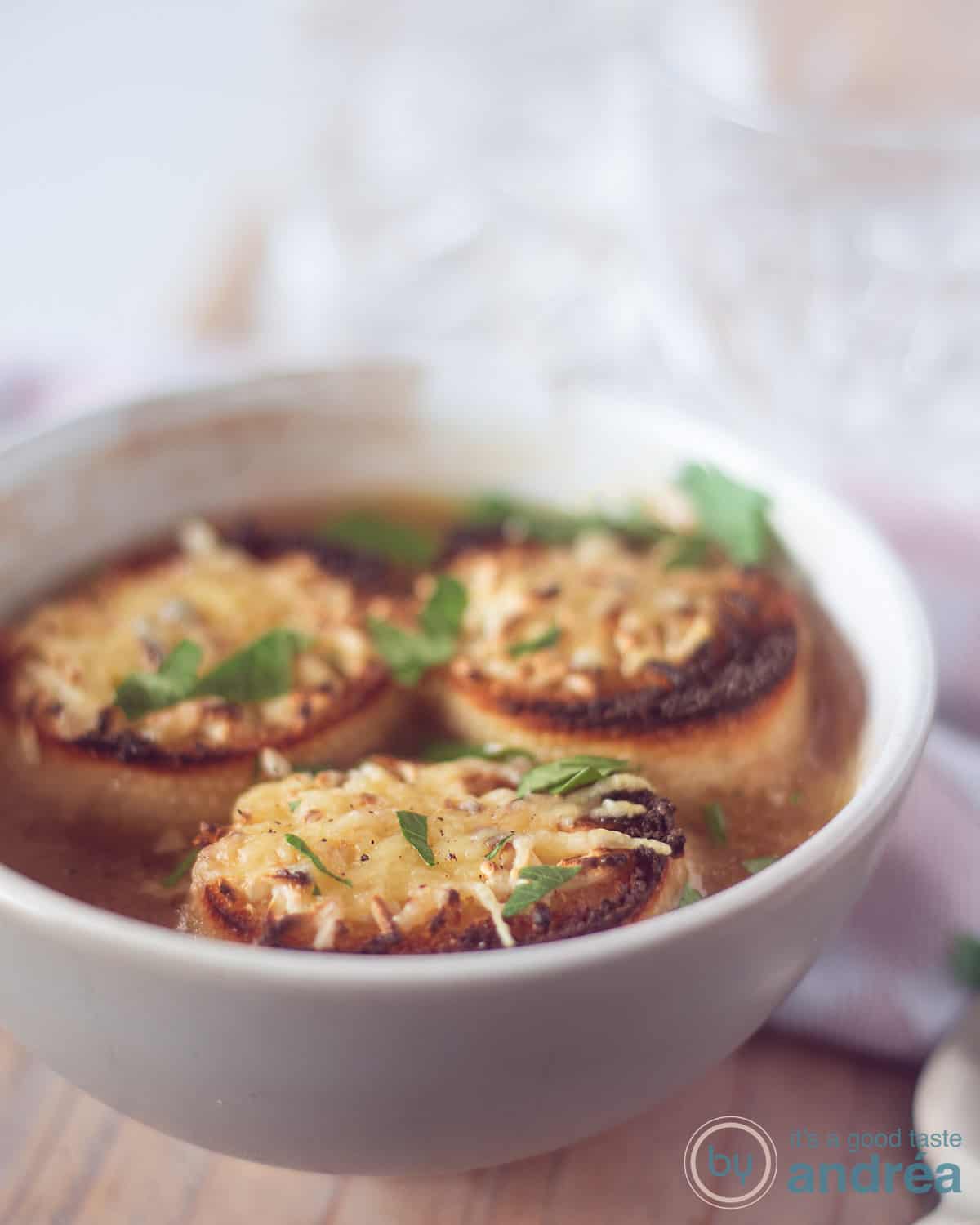 A height photo of a gray bowl of beer onion soup with three baguettes floating on top with gratinated cheese. On a wooden plank.