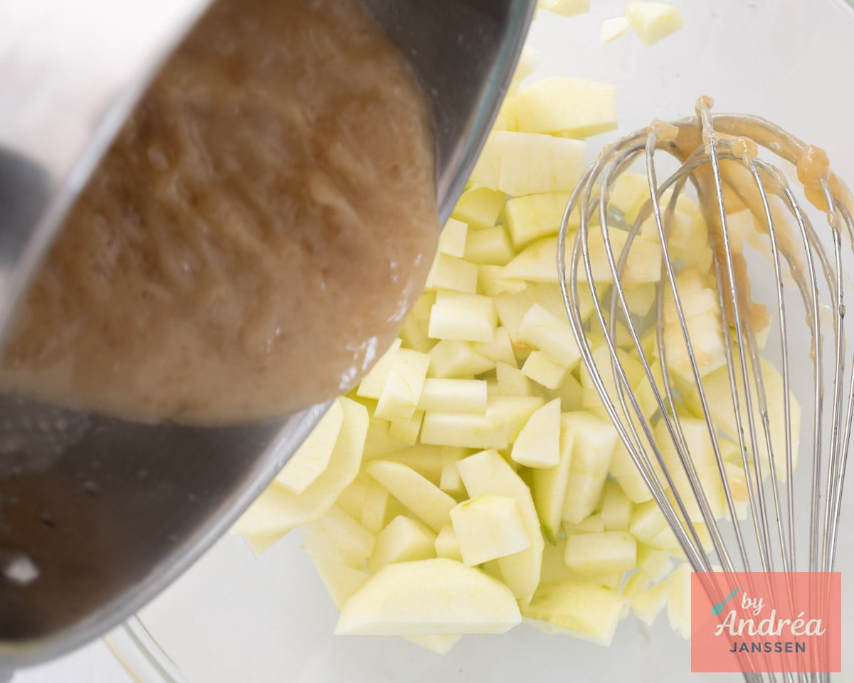 A saucepan of light brown sauce is tilted over a glass bowl of peeled, chopped apples with a whisk inside.
