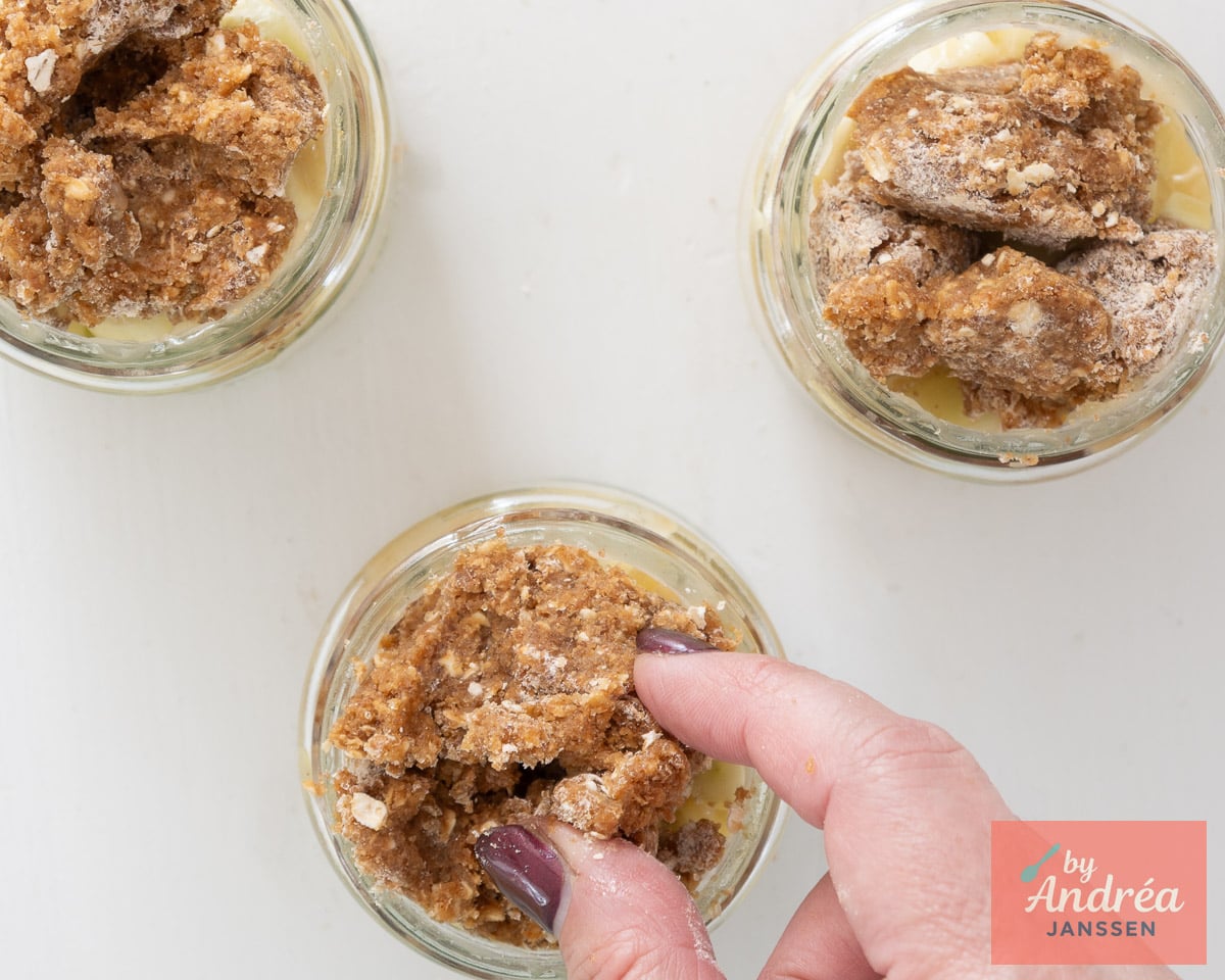 A hand adds oat crumble topping to apple filling in three glass jars.