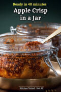 Glass jar of apple crisp with an oat topping and a gold spoon on a brass plate.