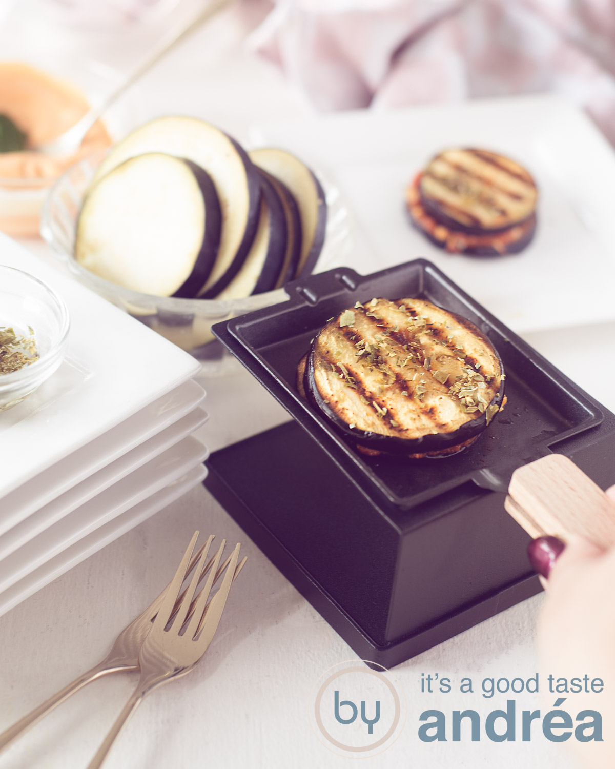 A black raclette dish with eggplant melt. A hand holding hte dish. Some eggplants above. A white background