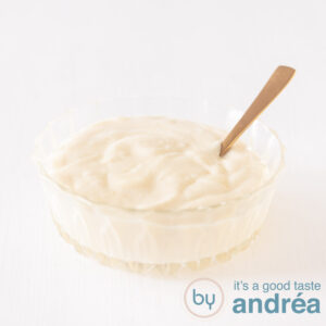 A glass bowl filled with homemade mayonnaise on a white background. A golden spoon stands in the sauce