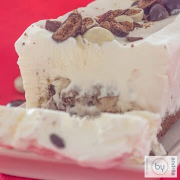 A square photo of an ice cream cake with chocolate gingerbread nuts. A slice cut off.