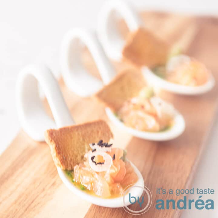 A wooden board with three white spoons filled with salmon, arugula oil and a piece of toast, angled