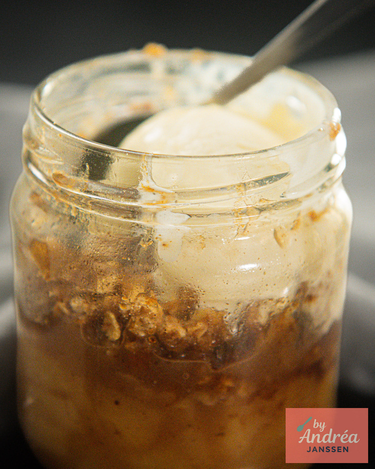 Close-up of apple crumble in a glass jar topped with a scoop of melting ice cream, with a spoon tucked inside.
