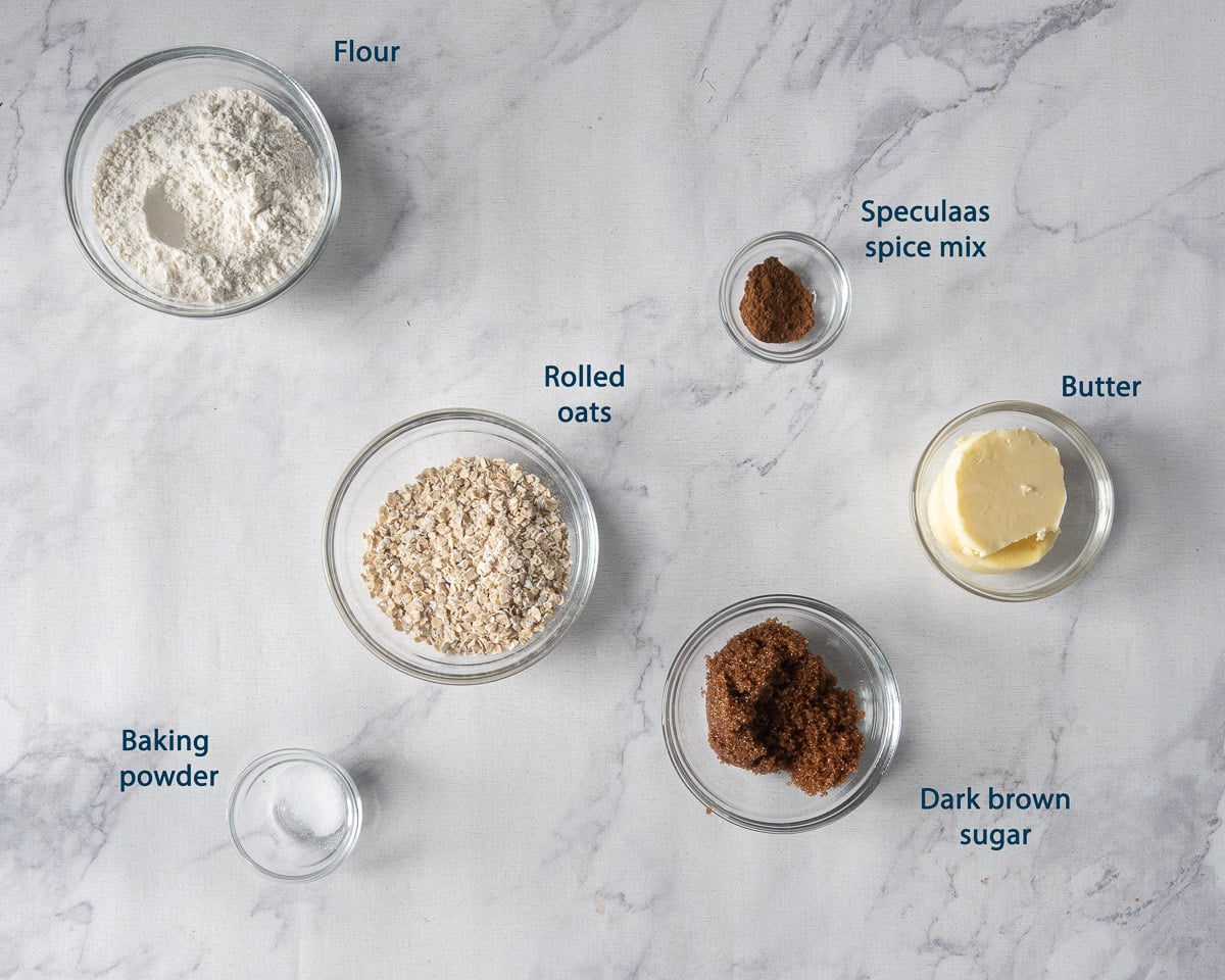 Bowls of flour, rolled oats, speculaas spice mix, butter, dark brown sugar, and baking powder with English labels.