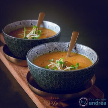 A square photo with two bowls on a gold platter on a wooden board. In the bowls Oriental pumpkin soup with cress.