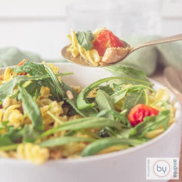 A square photo showing part of a white bowl filled with fusilli, arugula, tomatoes and pesto.