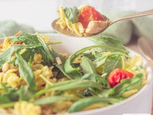 A square photo showing part of a white bowl filled with fusilli, arugula, tomatoes and pesto.