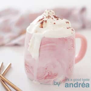 A square photo of a pink mug of Baileys with hot chocolate and a whipped cream topping. A white background, in the background a pink and white tea towel. In the front left two gold straws