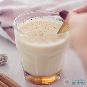 A square photo with a glass in the middle filled with chai tea with milk and honey. A golden spoon stirs the glass.