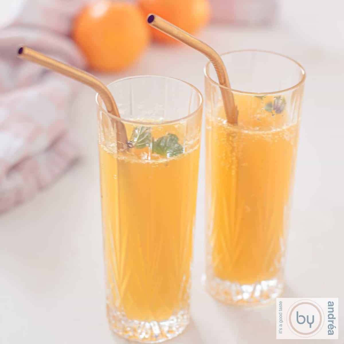 A square photo of two glasses with mandarin champagne cocktail a golden straw. Two mandarins in the background.