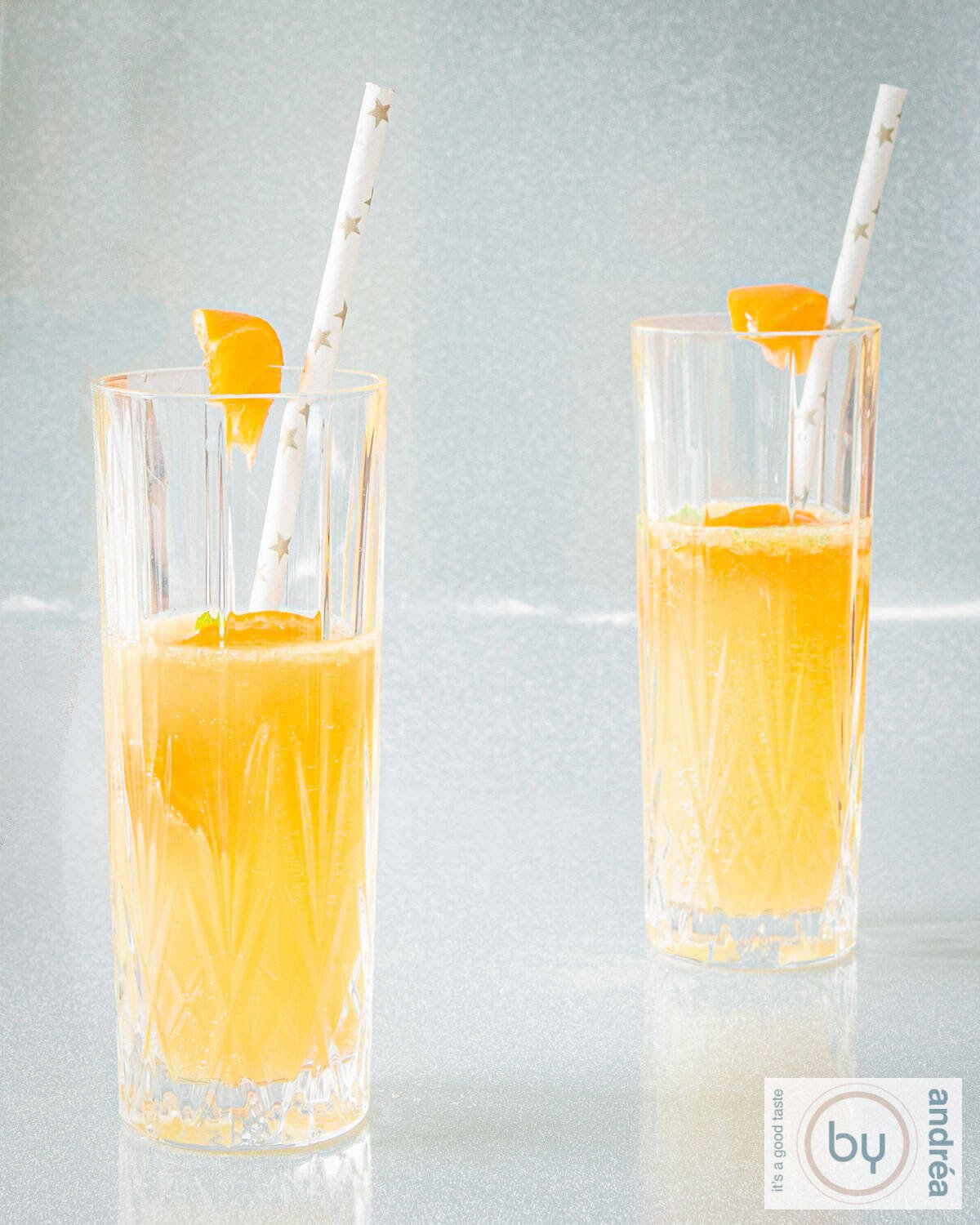 A height photo of two glasses filled with a cocktail of mandarin, champagne and cointreau on a silver background.