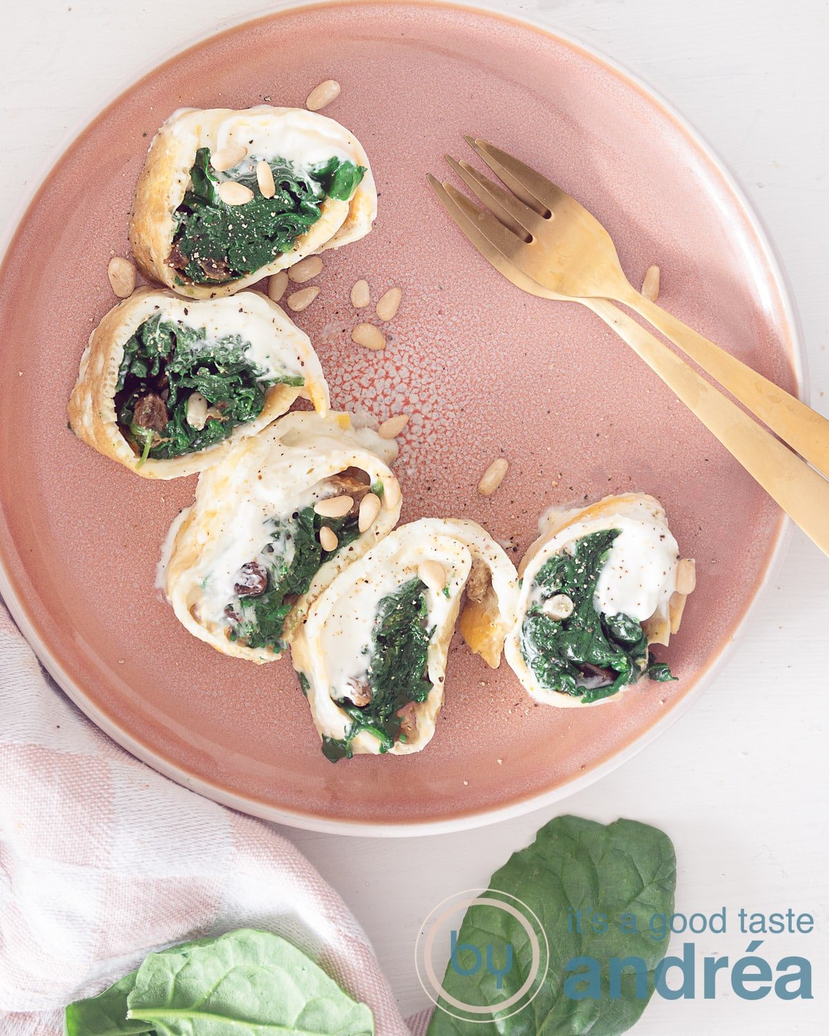 A photo from above with a pink plate at the top with a half moon of spinach sour cream wraps. Next to it two golden forks. Spinach leaves at the bottom