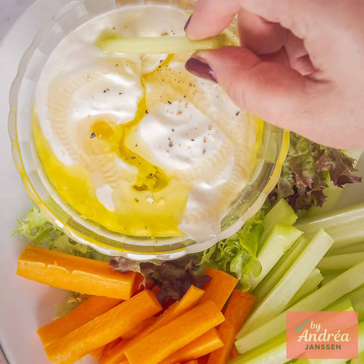 A square photo of creamy whipped feta dip with olive oil and vegetable sticks around it. A hand dips vegetables in the feta sauce.