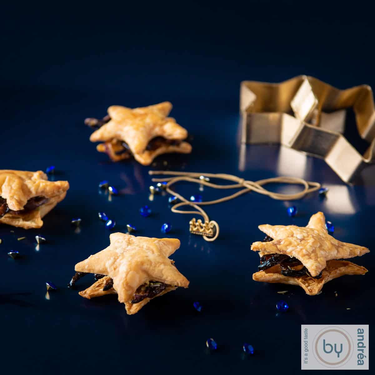 Een vierkante foto met een blauwe glanzende ondergrond met daarop bladerdeeg sterren gevuld met gekarameliseerde uien en tijm. Gouden accesoires eromheen