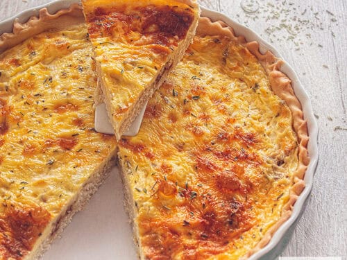 A square photo of a gray dish filled with goat cheese and shallot quiche. A wedge has been cut out and is resting on a white cake server.