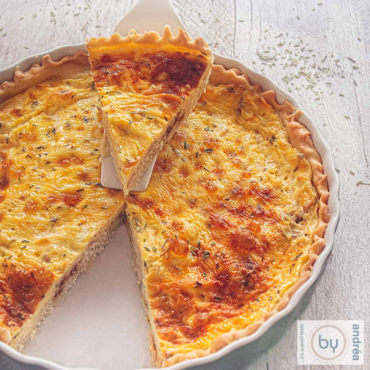 A square photo of a gray dish filled with goat cheese and shallot quiche. A wedge has been cut out and is resting on a white cake server.