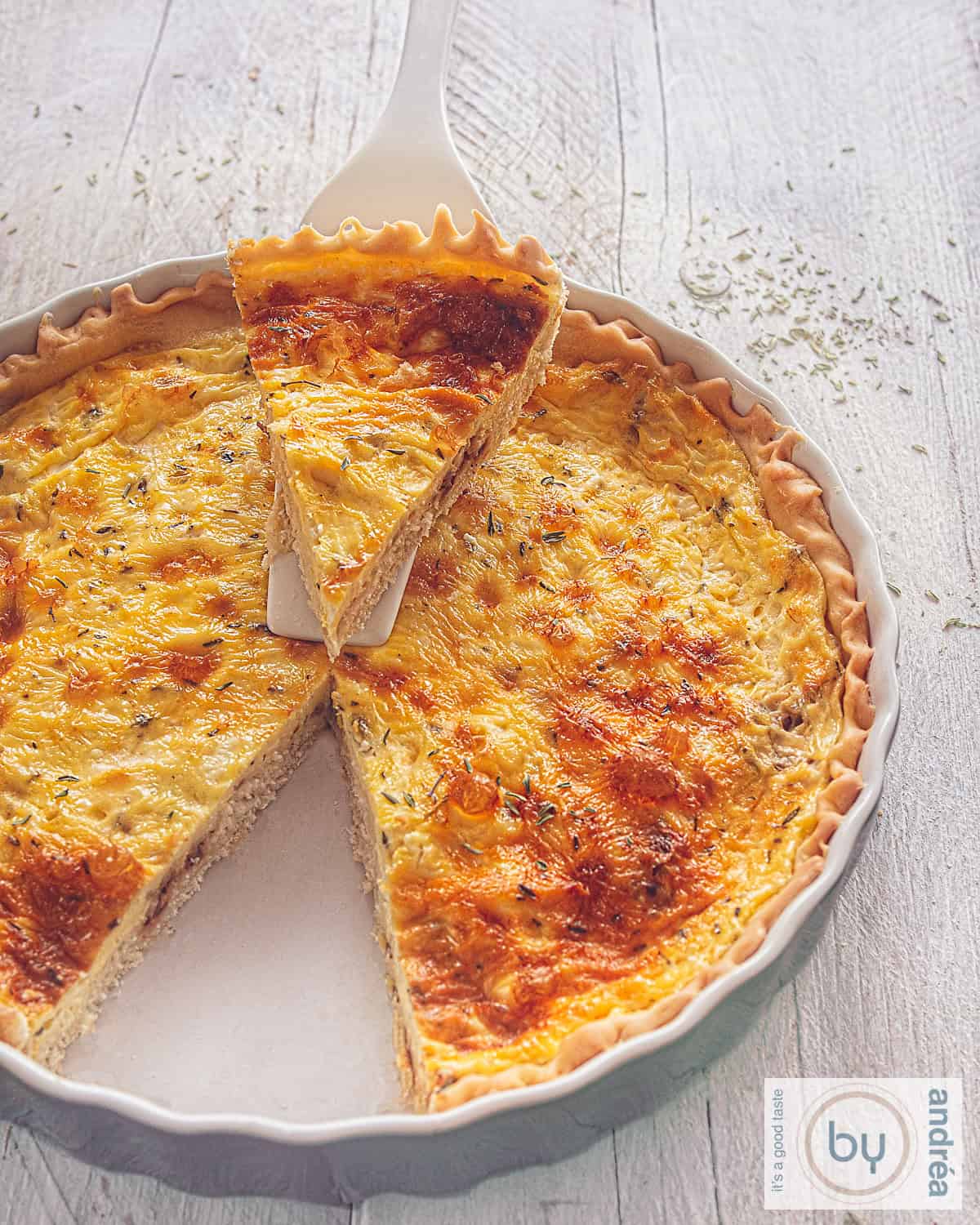 A height photo of a gray quiche dish filled with goat cheese, egg, thyme and shallot quiche from which a point has been cut out which lies on a cake server on top