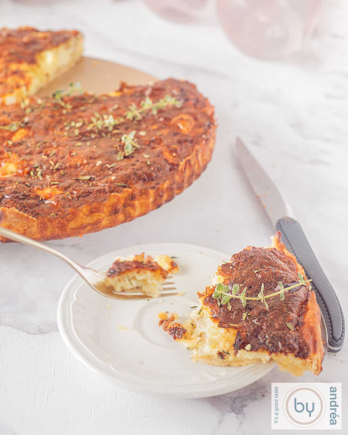 A height shot of a white plate with a slice of goat cheese quiche and the rest of the quiche in the background.