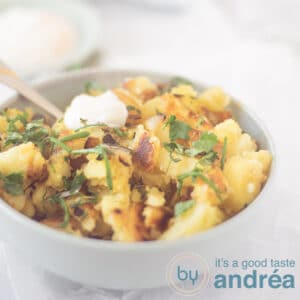 A square photo of a blue bowl filled with potatoes with curry and herbs with a dollop of garlic yogurt sauce