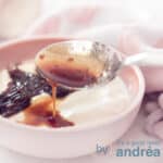 A square photo with part of a pink dish with soaked plum compote on Greek yogurt. A pink and white checkered tea towel lies diagonally above it.