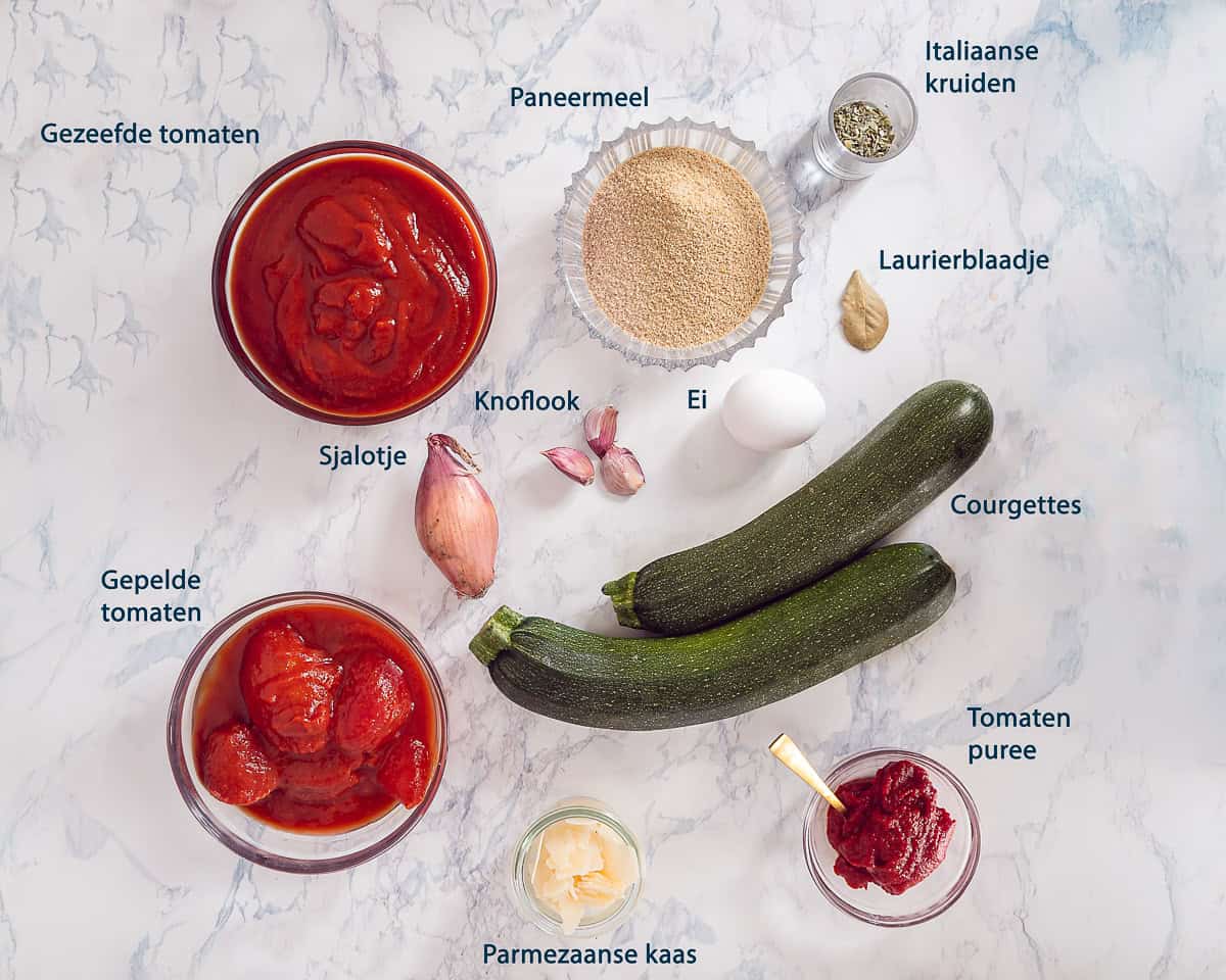 Ingredienten Courgetteballetjes In Tomatensaus uit de Oven op een wit marmeren ondergrond met tekstbeschrijving.