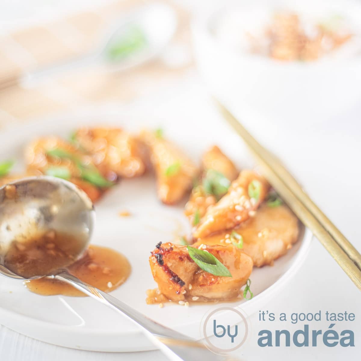A white plate with pieces of honey sesame baked chicken, garnished with spring onions. A spoon with extra sauce in front. A white background with chopsticks, exttra chicken and a bamboo mag