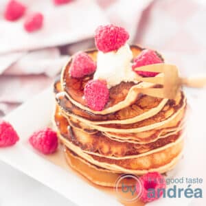 A square photo with a stack of pancakes, raspberries on top and around it and a dot of crème frache. In the background a pink white cloth