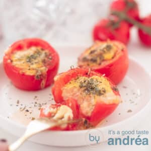 A square photo with tomatoes stuffed with egg, cheese and herbs. A fork pricks a piece off a glass dish. In the background a bunch of tomatoes