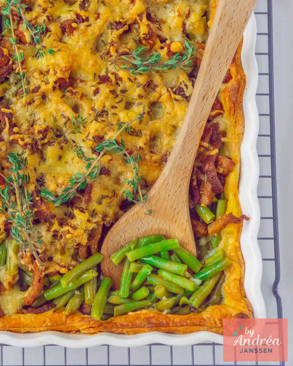 Een foto van bovenaf van een shoarma ovenschotel met boontjes, kaas, en shoarmavlees. Een houten lepel neemt een schep.