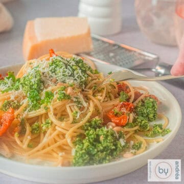 A square photo of a white plate with spaghetti, kale pesto and sun-dried tomatoes. A fork is poking the plate.