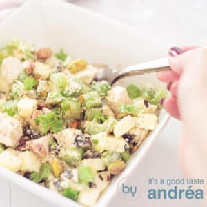 A square photo of a square white bowl filled with Waldorf salad. A hand is holding a spoon grabbing some salad. On a white background