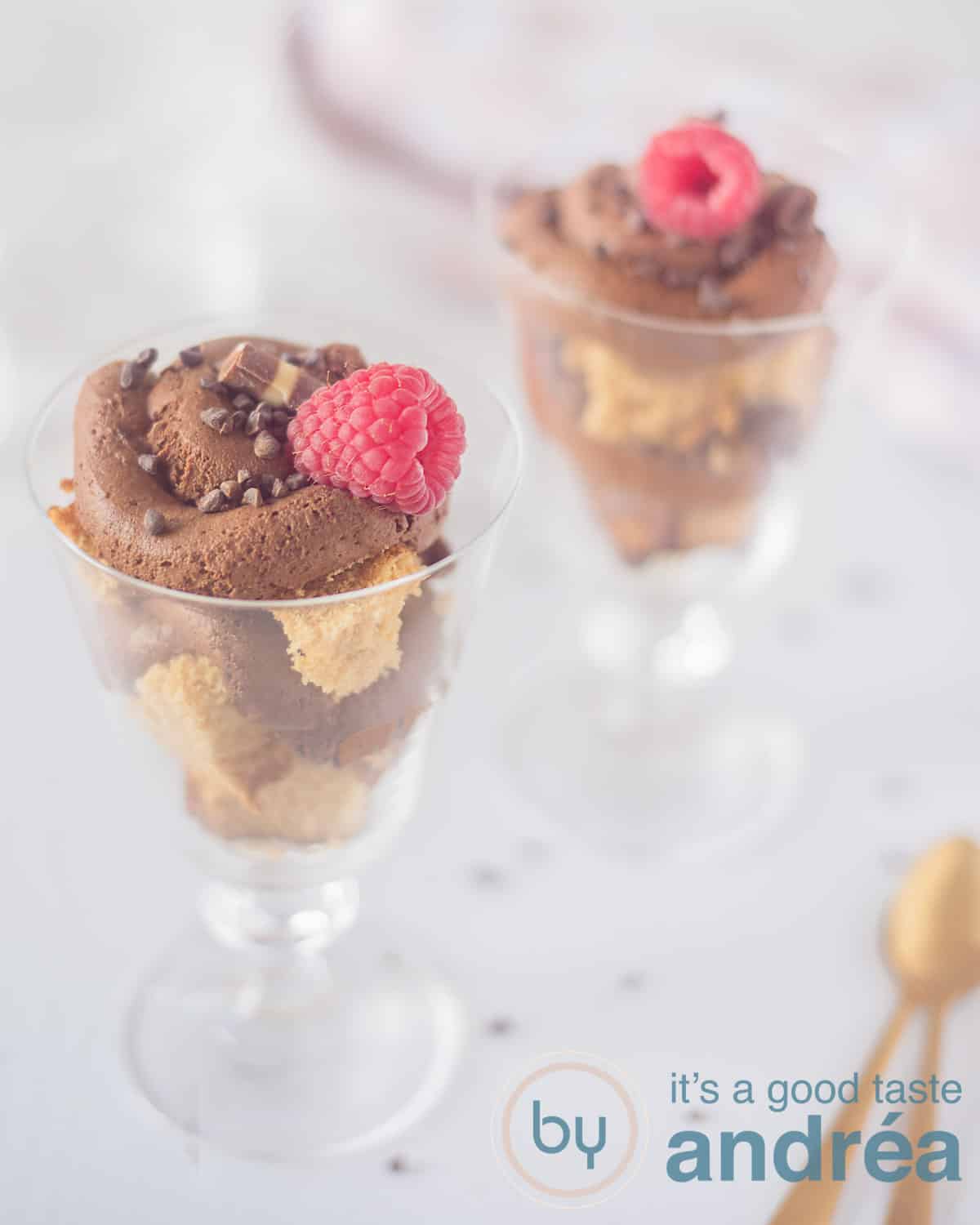 A height photo with two glass coupes filled with layers of cake alternated with chocolate mousse and top with a raspberry and chocolate. A white background. Bottom right two golden spoons.