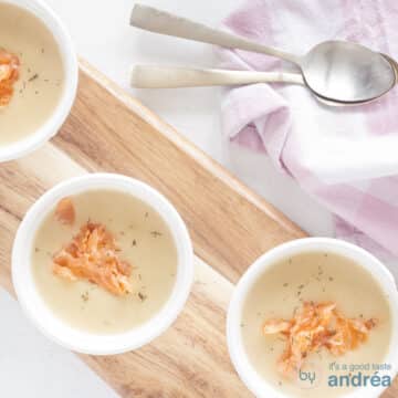 A square photo from above with three white cups filled with thickened queen soup with a garnish of smoked salmon and dill. At the top right a pink white tea towel and two gold spoons.