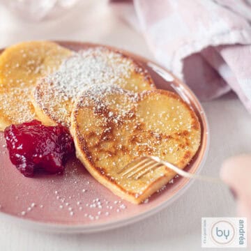 A square photo with a pink plate with heart-shaped pancakes with powdered sugar and strawberry jam. A fork is poking a pancake.