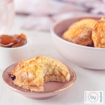 A square picture of a pink plate with a scone, a bite taken out of it. In the back a bowl with more scones and some orange jelly.