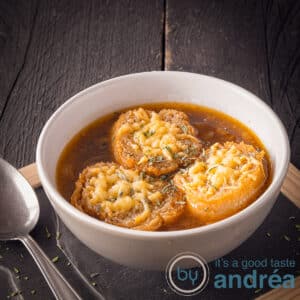 A square photo with a black background on top of a black plate with a bowl filled with onion soup and baguette au gratin with cheese. A spoon on the left