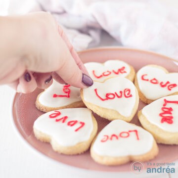 A square photo with a pink plate with heart cookies with icing and text in red. A hand grabs a cookie.