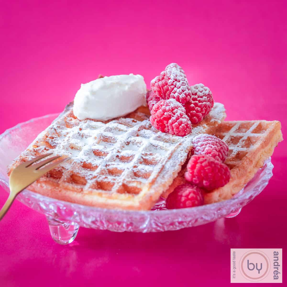 A square photo with a glass bowl with 2 waffles, raspberries and mascarpone. A pink background.
