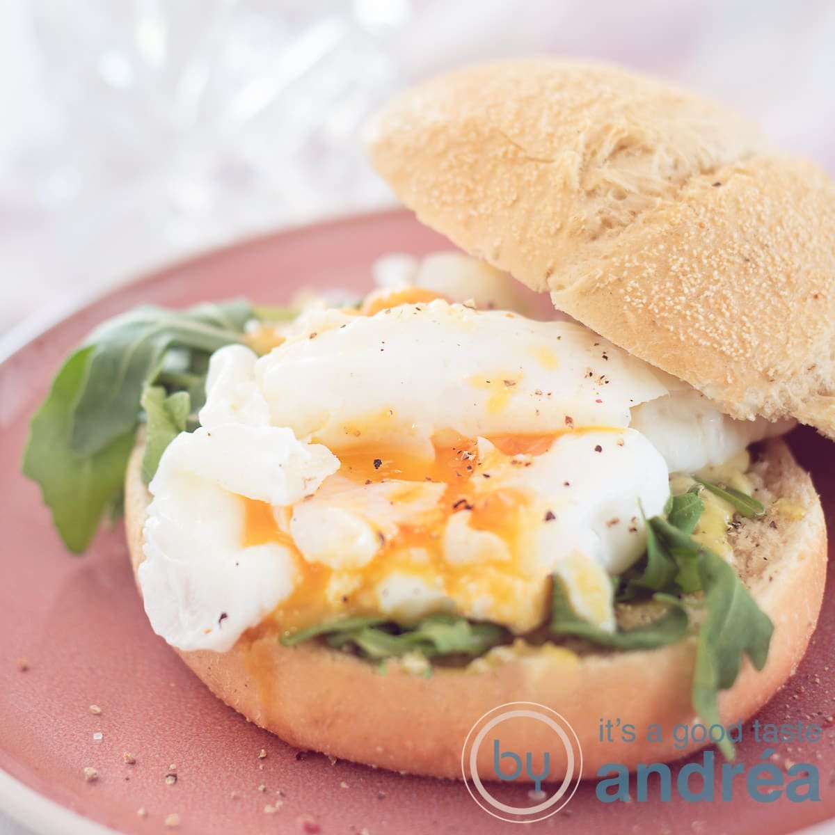 A square photo with a pink plate with a crispy bun with pesto mayonnaise, arugula and poached egg on a white background.