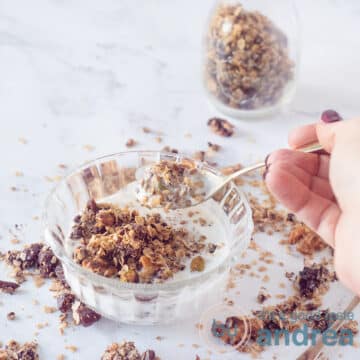 A square photo with a glass bowl with yogurt and chocolate muesli. A golden spoon takes a bite on a marble white background. In the background a glass jar with more chocolate nut granola.