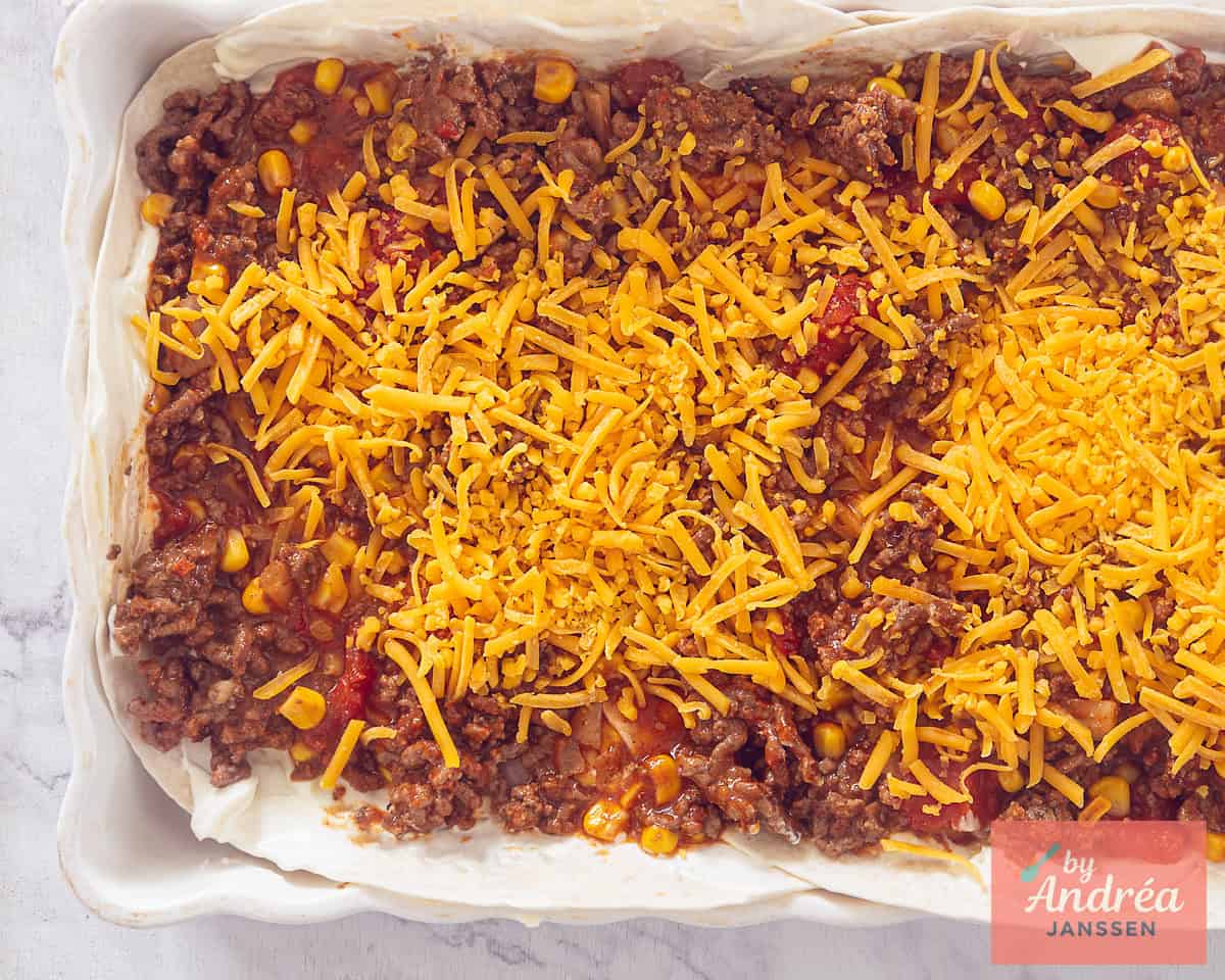 Een ovenschaal die wordt gevuld met gehaktsaus, tortilla's en bestrooid met kaas