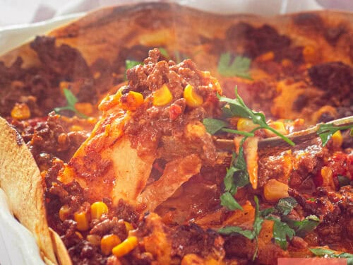A square photo showing part of a white baking dish with minced meat, tomato sauce, tortillas, corn and cheese.