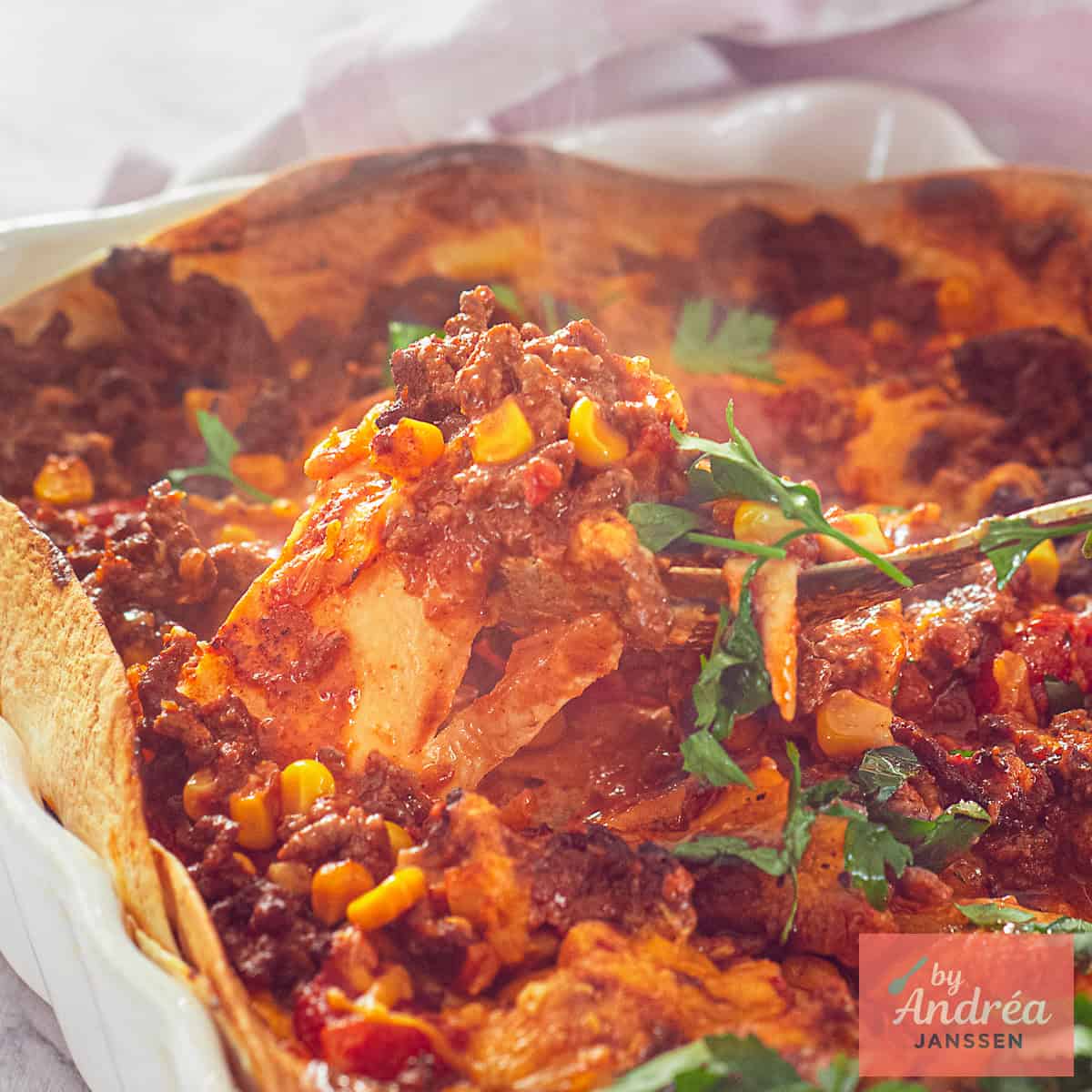 A square photo showing part of a white baking dish with minced meat, tomato sauce, tortillas, corn and cheese.