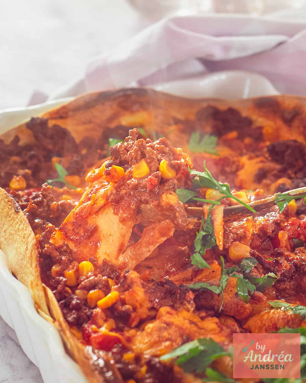 Een deel van een ovenschaal gevuld met Mexicaanse tortilla, tomatensaus, gehakt en kaas. Een lepel neemt een schep.