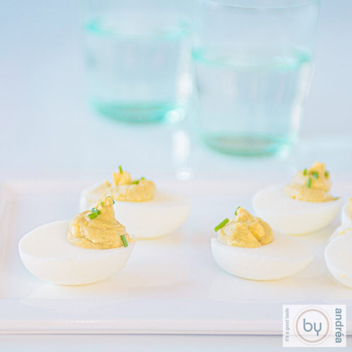 A square picture with a white plate with avocado curry deviled eggs. Two glasses in the background