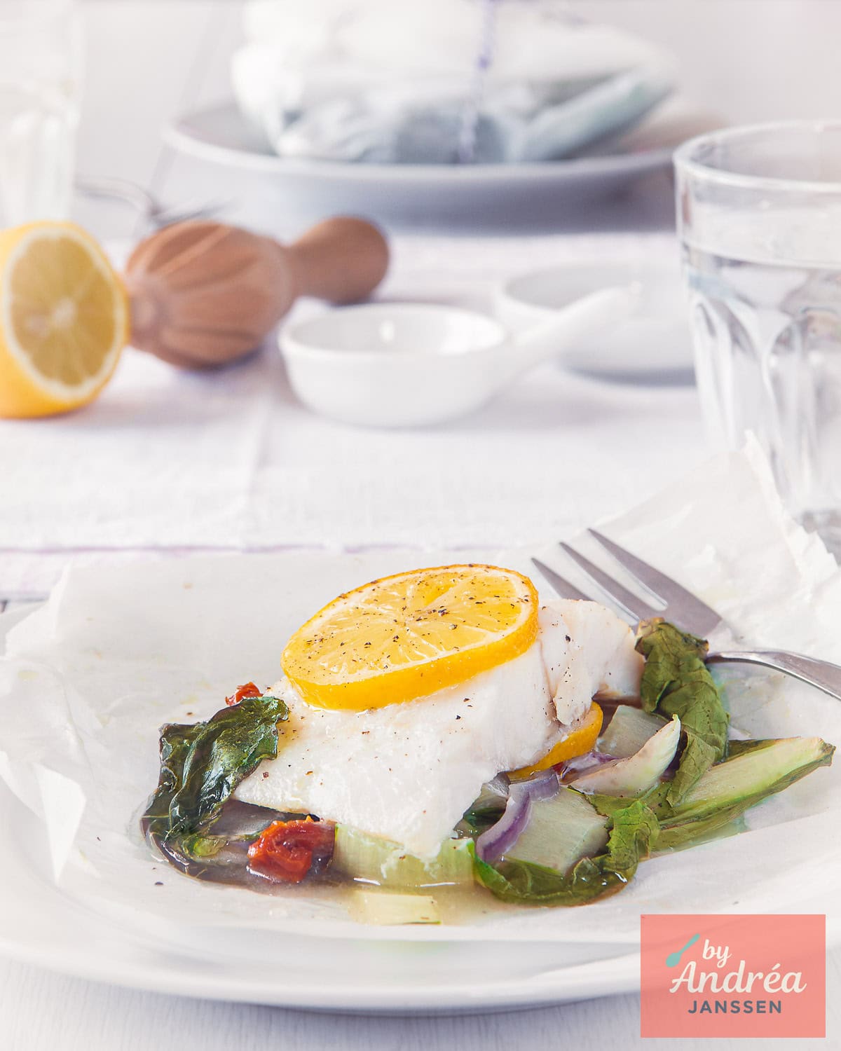 In the foreground, a plate with a fish parcel with bok choy, red onion, and lemon. In the background, cooking utensils and tableware.