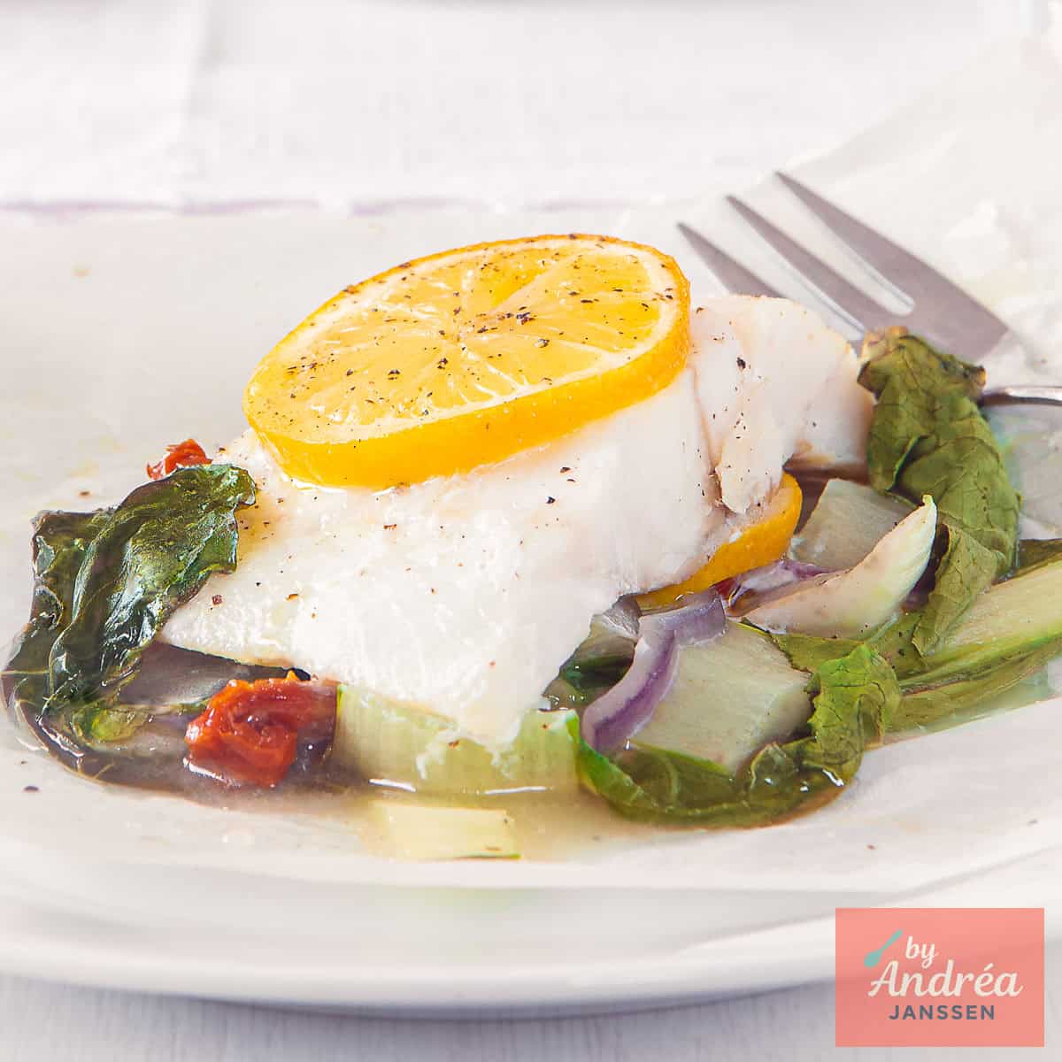 A square photo of a plate with bok choy, sun-dried tomatoes and red onion, topped with fish and lemon.