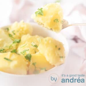 A square shot of a white bowl filled with potatoes with parsley and chives. A spoon takes out a potato on a white background